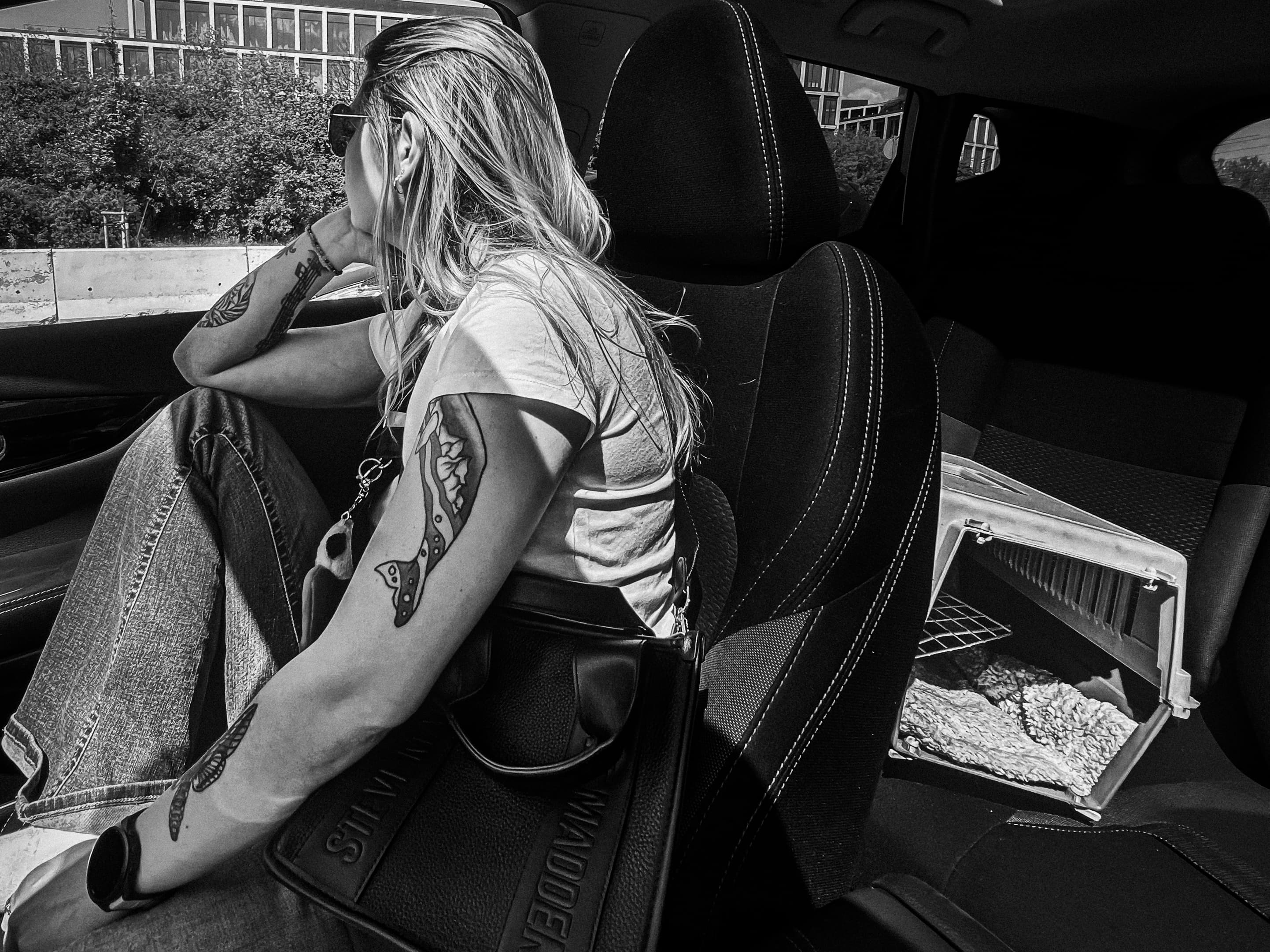 Black-and-white photograph of a woman sitting in a car, looking out the window, with a pet carrier on the seat beside her.